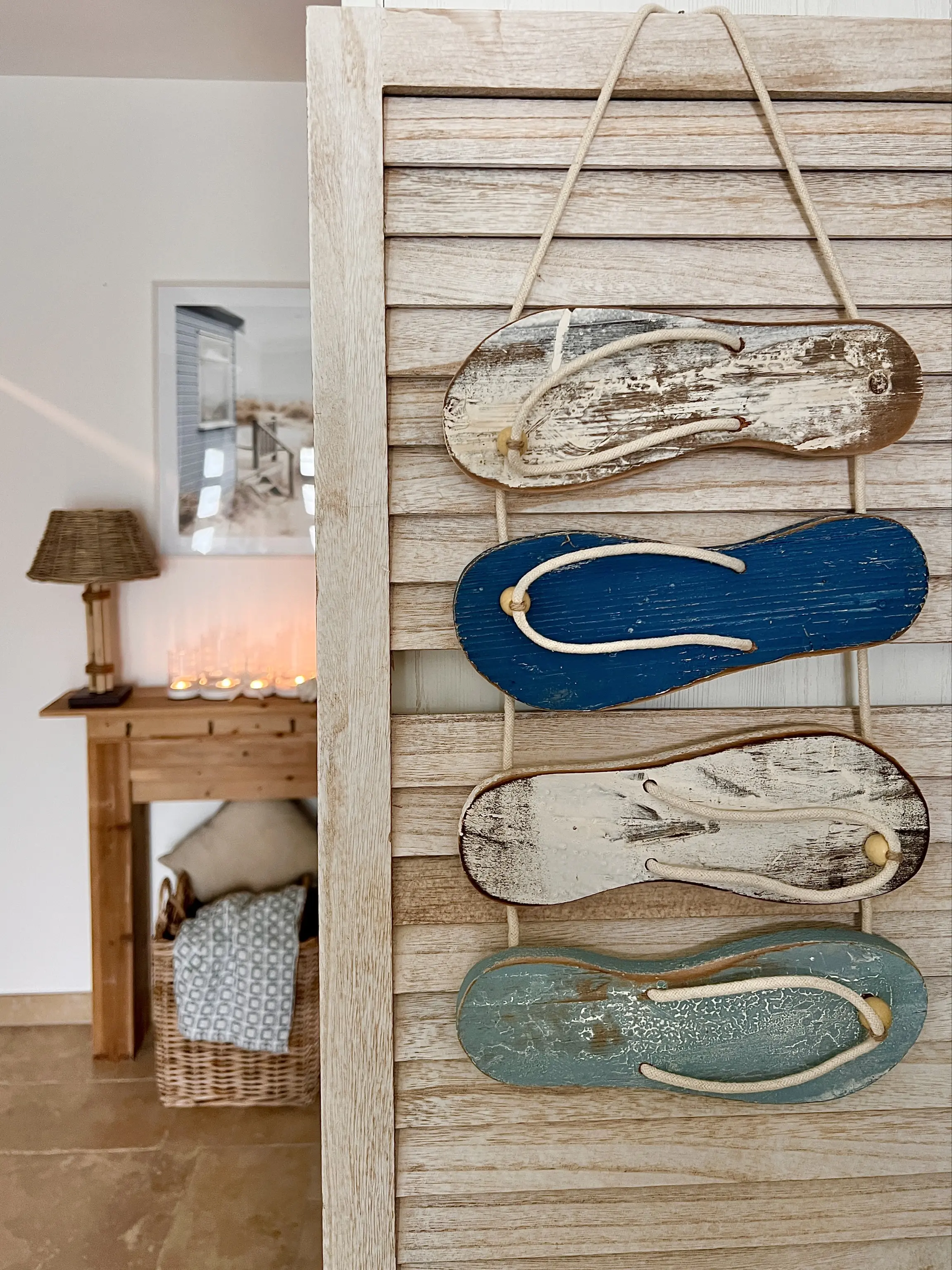 Dekoratives Holzpaneel mit aufgehängten Flipflops im Vintage‑Look in Blau- und Naturtönen, im Hintergrund Sideboard mit Kerzenlicht und maritimer Einrichtung