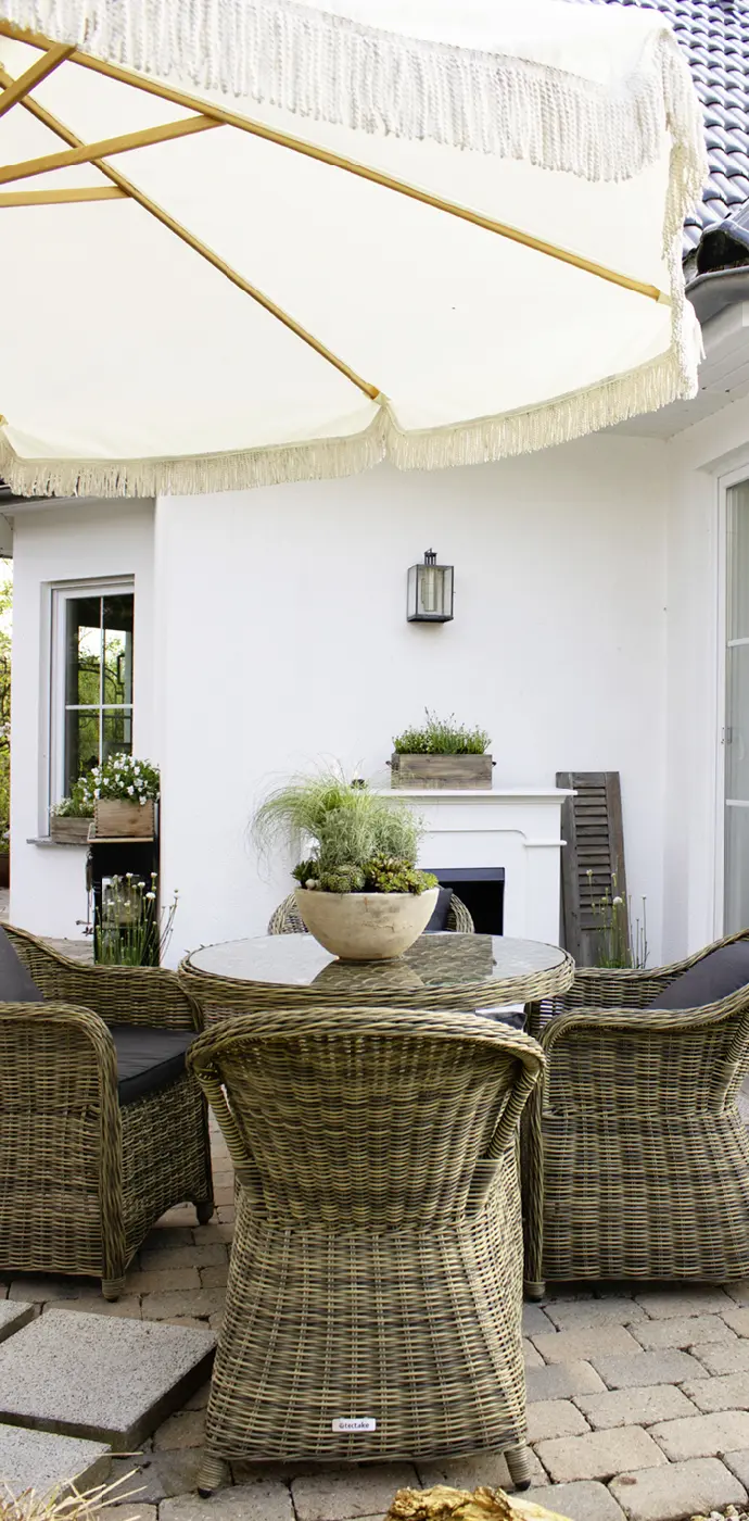Terrasse avec un coin salon rond en rotin, un parasol, des paniers fleuris et une cheminée d'extérieur devant la façade blanche de la maison