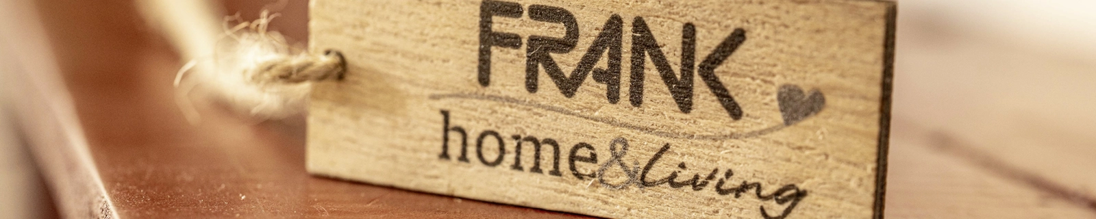Panneau en bois avec inscription gravée «FRANK home & living» et cordon de suspension sur une surface en bois