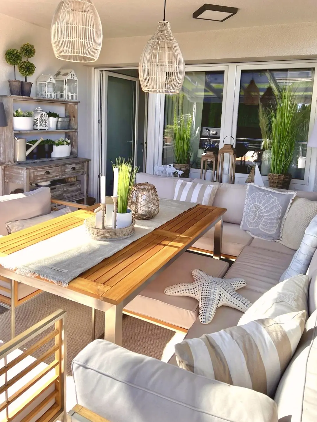 Gemütliche Terrassenlounge mit Ecksofa, Holztisch, maritimen Kissen, Pflanzen und Rattanlampen an der Decke