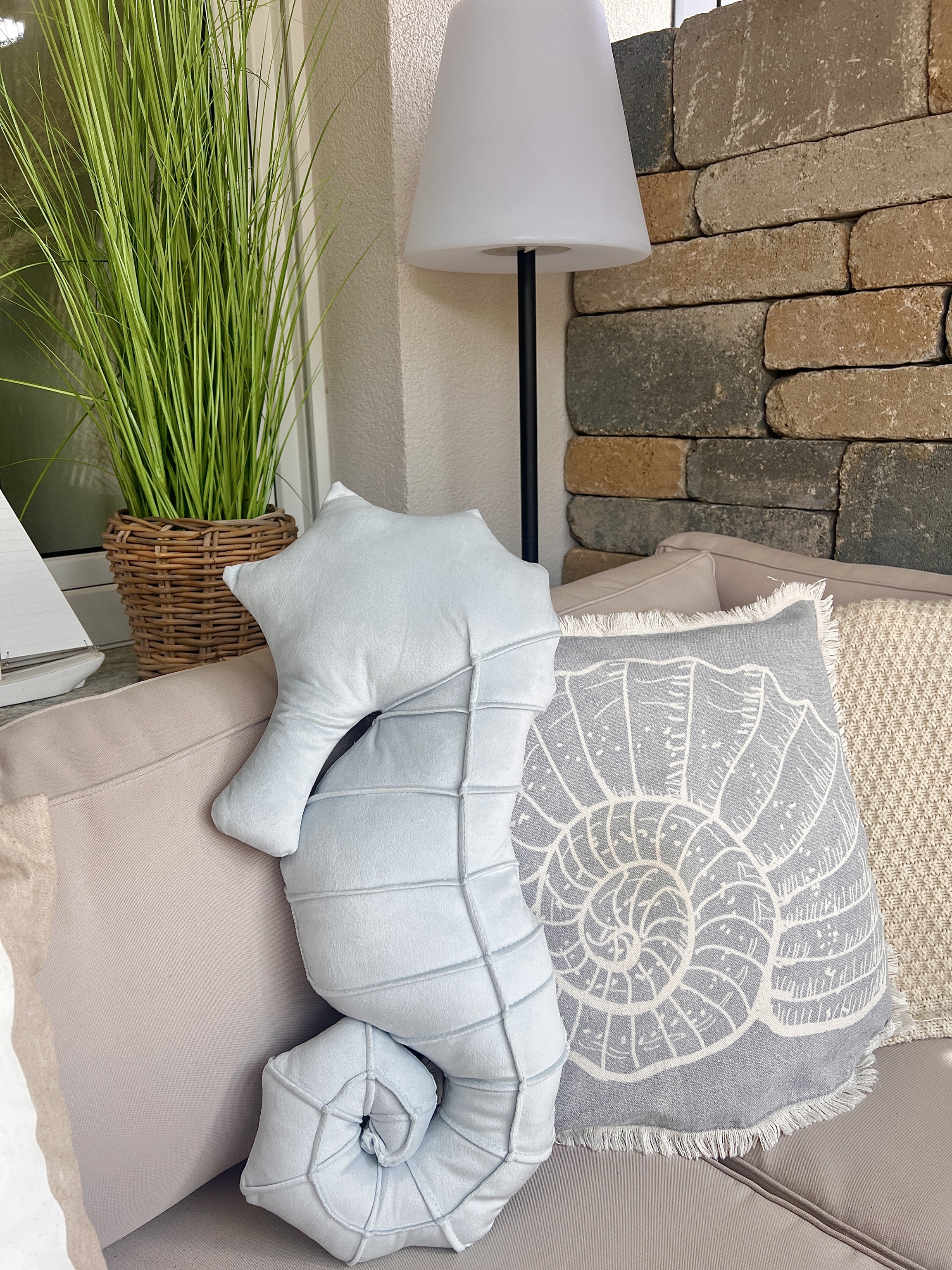 Zierkissen mit Muschelmotiv und Kissen in Seepferdchenform auf einer Outdoor‑Sitzbank neben Pflanze und Stehlampe