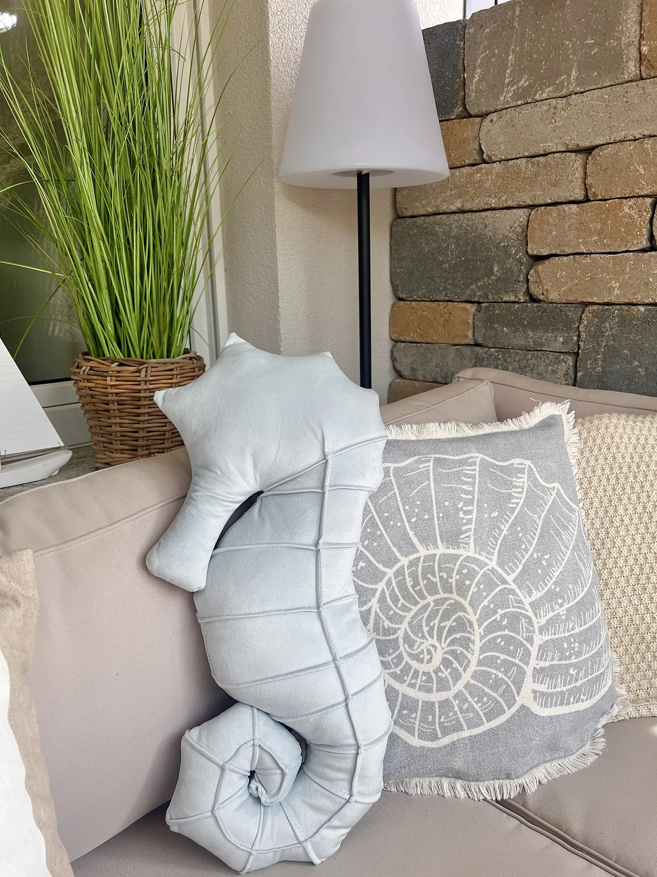Zierkissen mit Muschelmotiv und Kissen in Seepferdchenform auf einer Outdoor‑Sitzbank neben Pflanze und Stehlampe