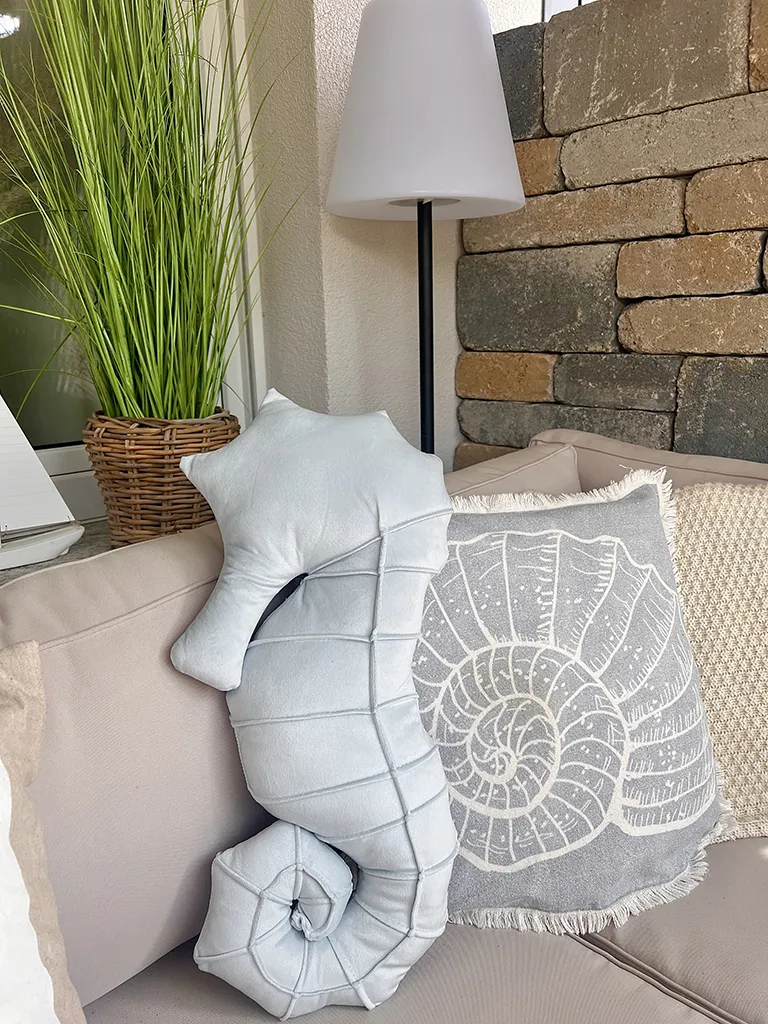 Zierkissen mit Muschelmotiv und Kissen in Seepferdchenform auf einer Outdoor‑Sitzbank neben Pflanze und Stehlampe