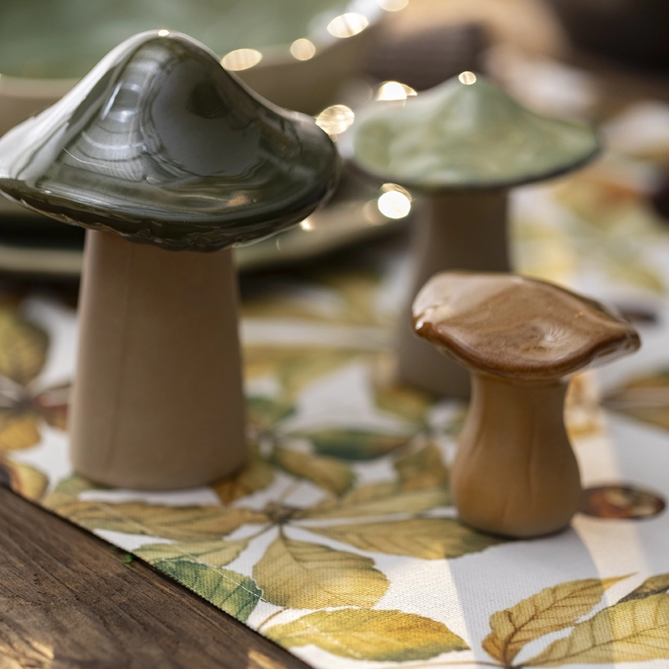 « Des champignons décoratifs en bois et en verre disposés sur un chemin de table orné d'un motif de feuilles d'automne, pour une décoration de table aux couleurs de l'automne »