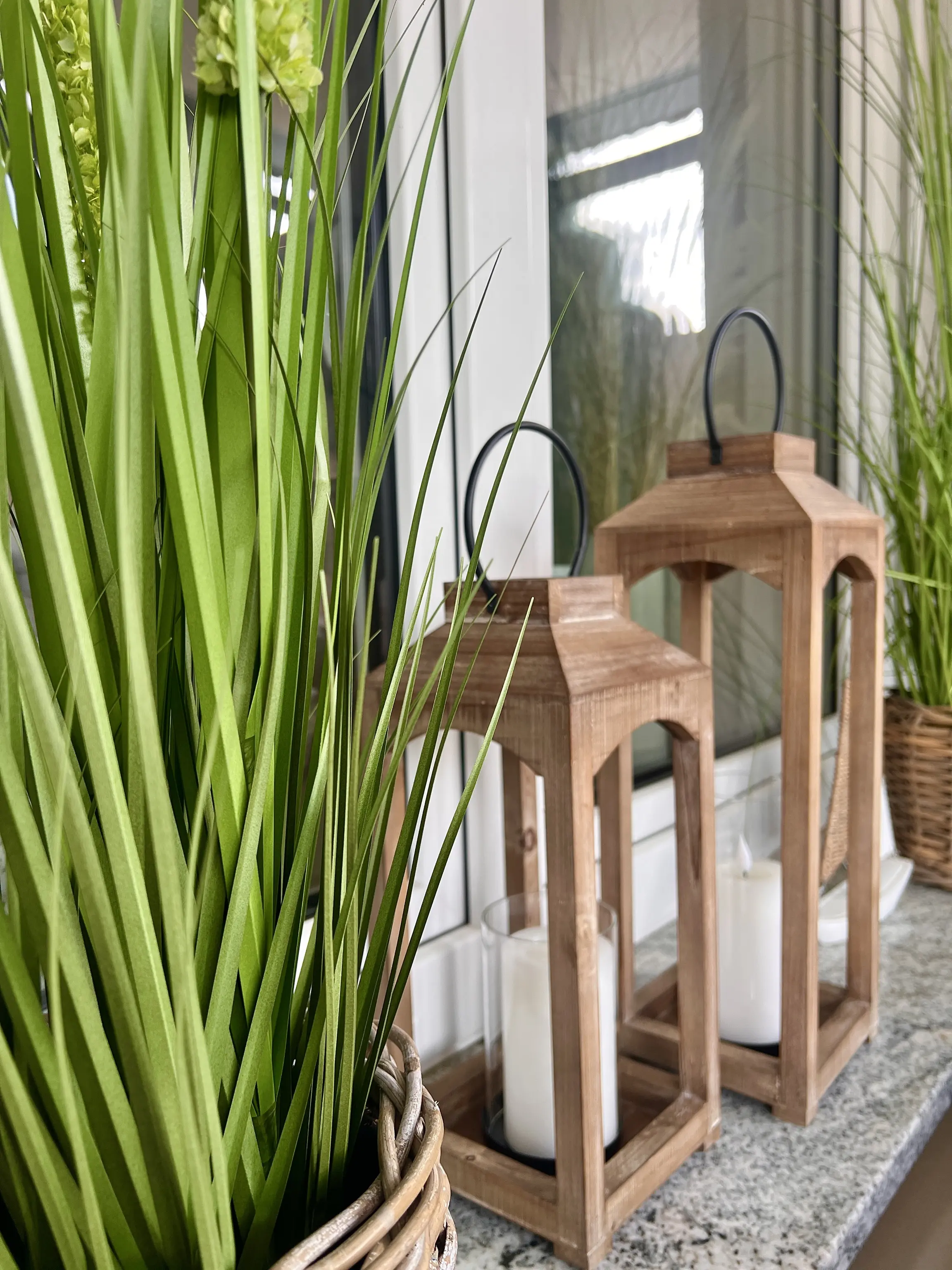 Holzlaternen mit weißen Kerzen auf einer Fensterbank neben hohen Ziergräsern in einem Korb