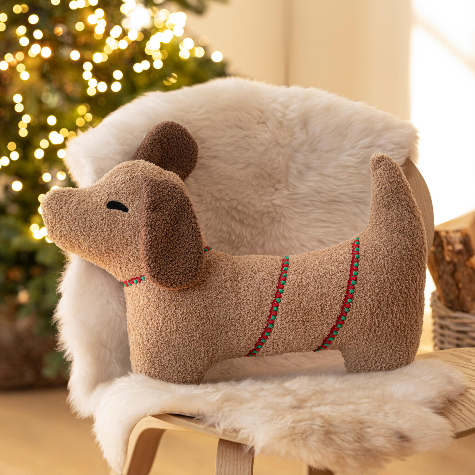 Coussin décoratif en forme de teckel posé sur un fauteuil recouvert de fausse fourrure blanche, avec en arrière-plan un sapin de Noël illuminé