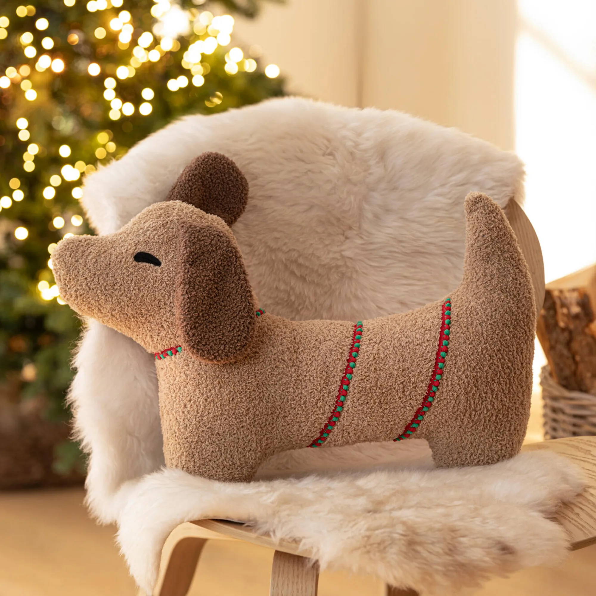 Coussin décoratif en forme de teckel posé sur un fauteuil recouvert de fausse fourrure blanche, avec en arrière-plan un sapin de Noël illuminé