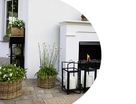 Décoration de terrasse avec une cheminée d'extérieur blanche, des lanternes en métal noir avec des bougies et des paniers tressés remplis de plantes