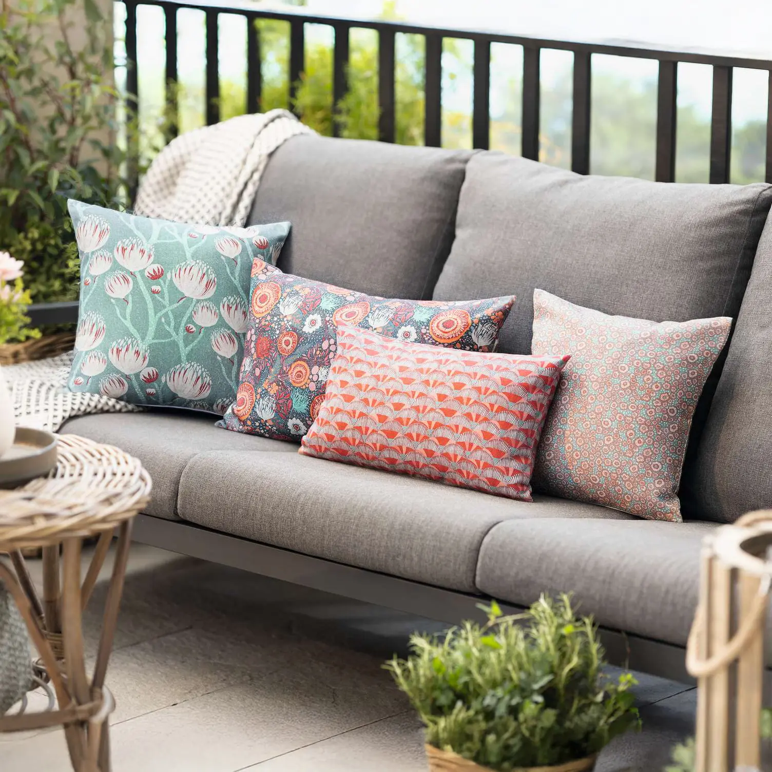 Canapé d'extérieur gris sur une terrasse avec des coussins décoratifs verts, orange et aux motifs colorés, accompagnés de plantes et de lanternes