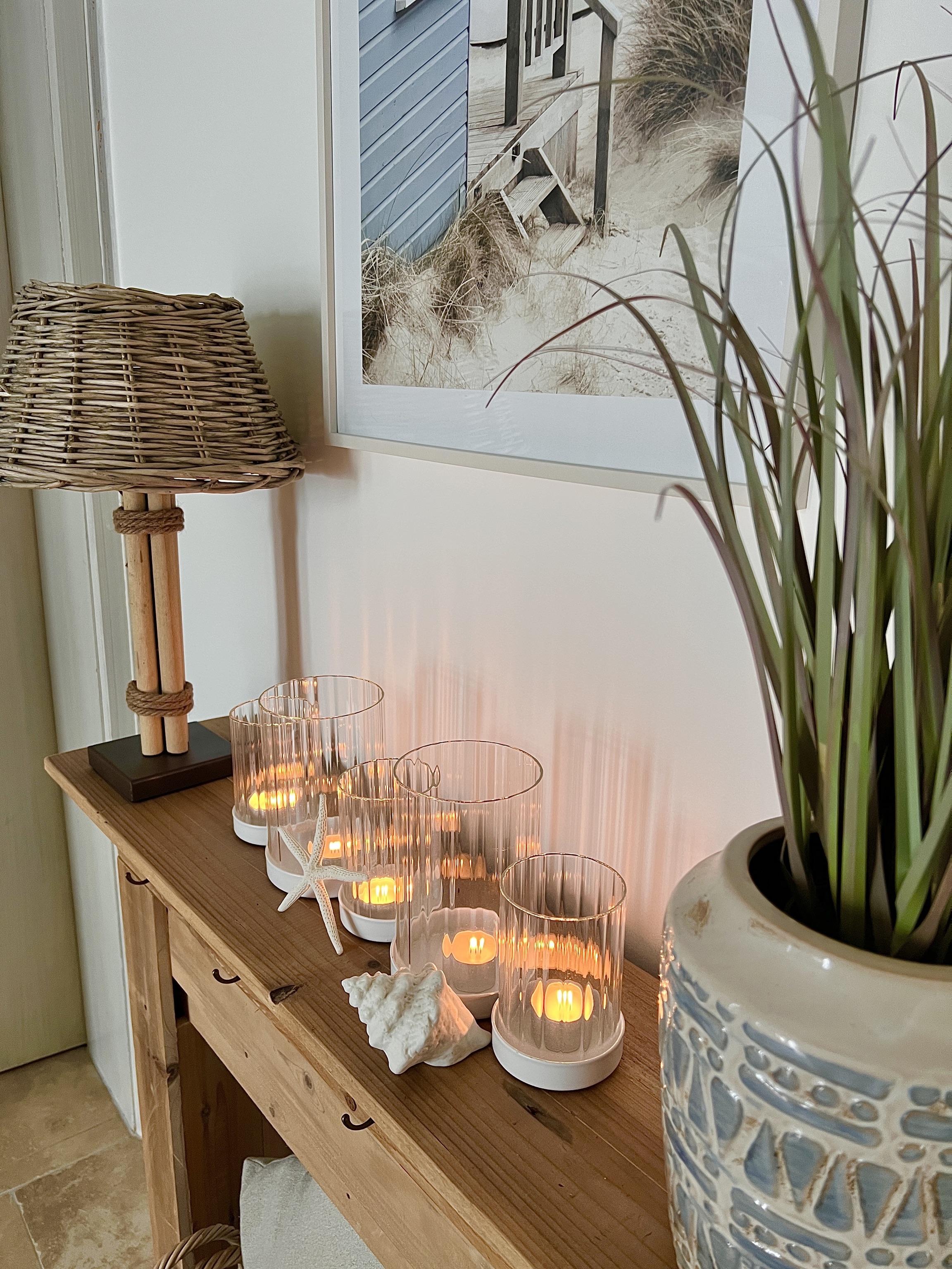Holz‑Sideboard mit mehreren Glaswindlichtern und brennenden Kerzen, dekoriert mit Muscheln, Stehlampe aus Rattan, Pflanze und Wandbild im maritimen Stil