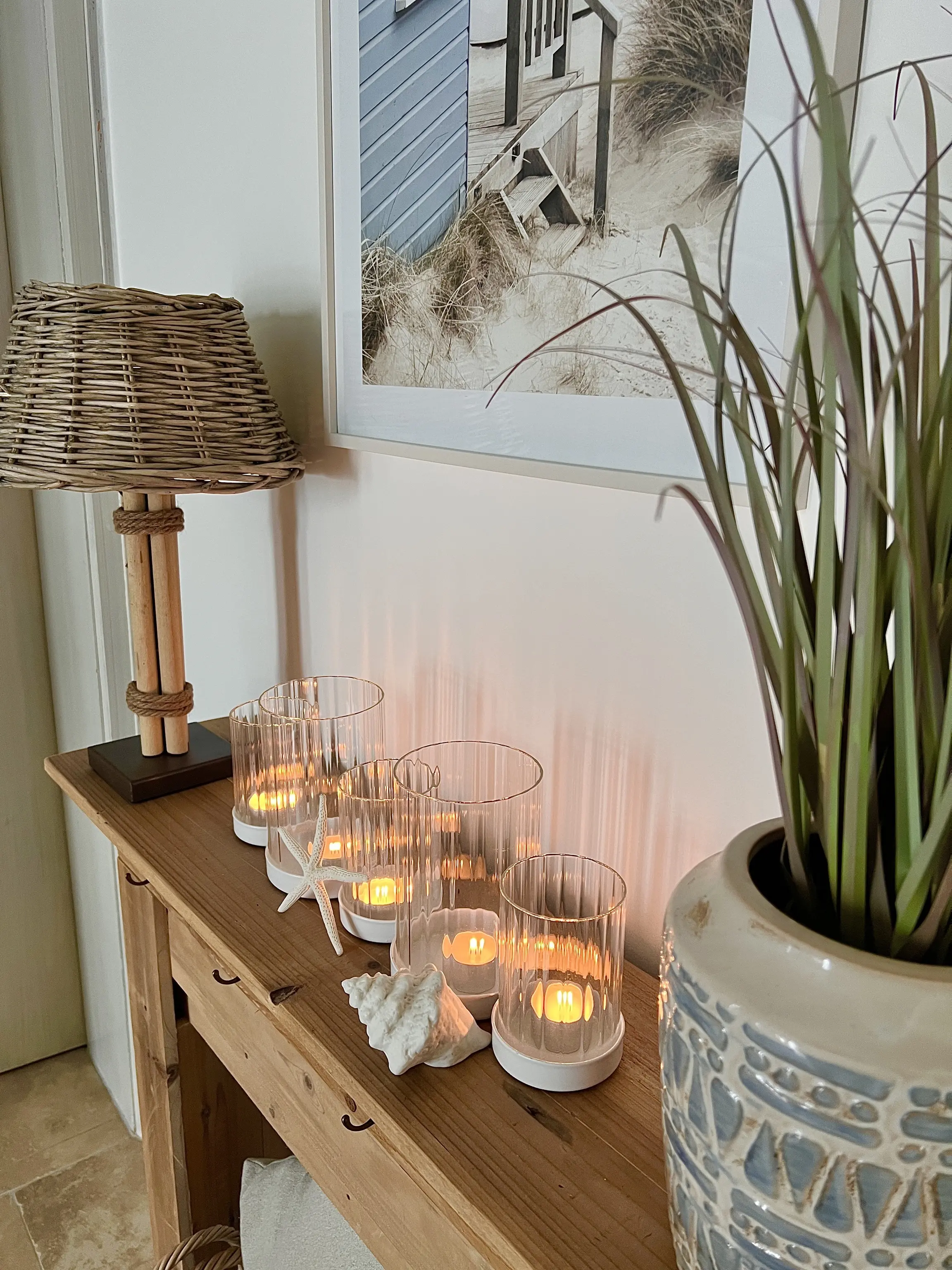 Holz‑Sideboard mit mehreren Glaswindlichtern und brennenden Kerzen, dekoriert mit Muscheln, Stehlampe aus Rattan, Pflanze und Wandbild im maritimen Stil