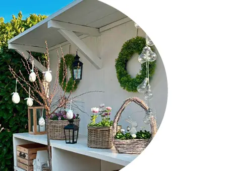 Décoration de Pâques printanière sur une étagère blanche avec des paniers remplis de plantes, des branches ornées d'œufs de Pâques, une couronne et des lanternes dans le jardin